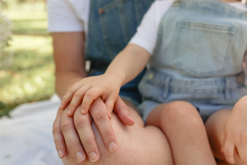 detail photo of a young girl placing her hand on top of her mother's hand that's resting on her mother's knee while the girl sits on her mother's lap