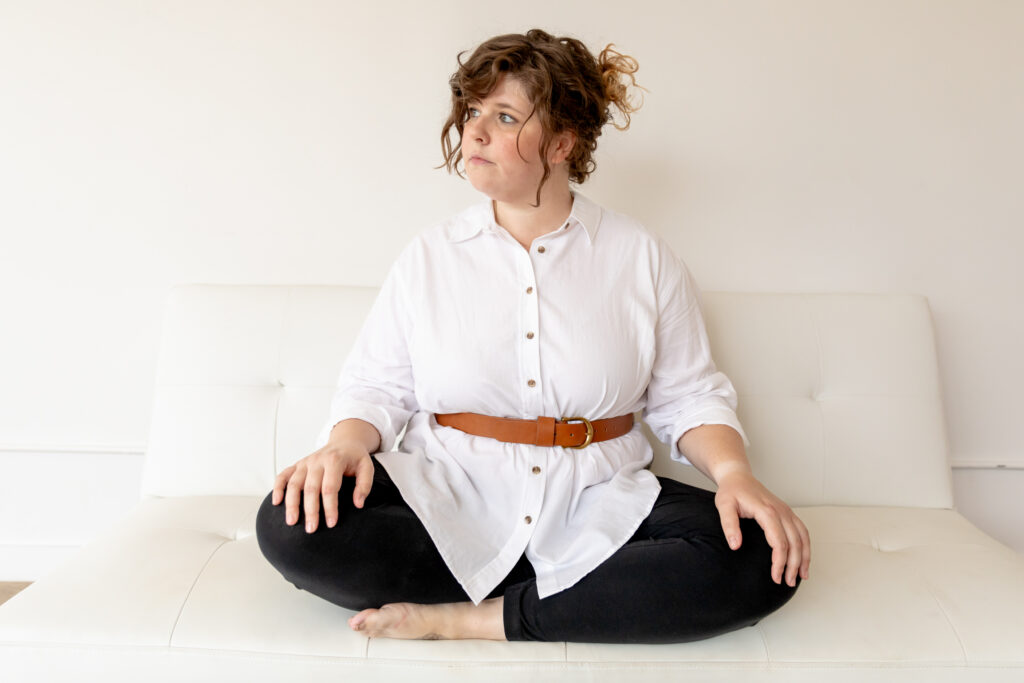 Emily of Emmy & Ollie Photography sits crosslegged on white couch and stares off to the left of frame at studiostudio in Ann Arbor Michigan