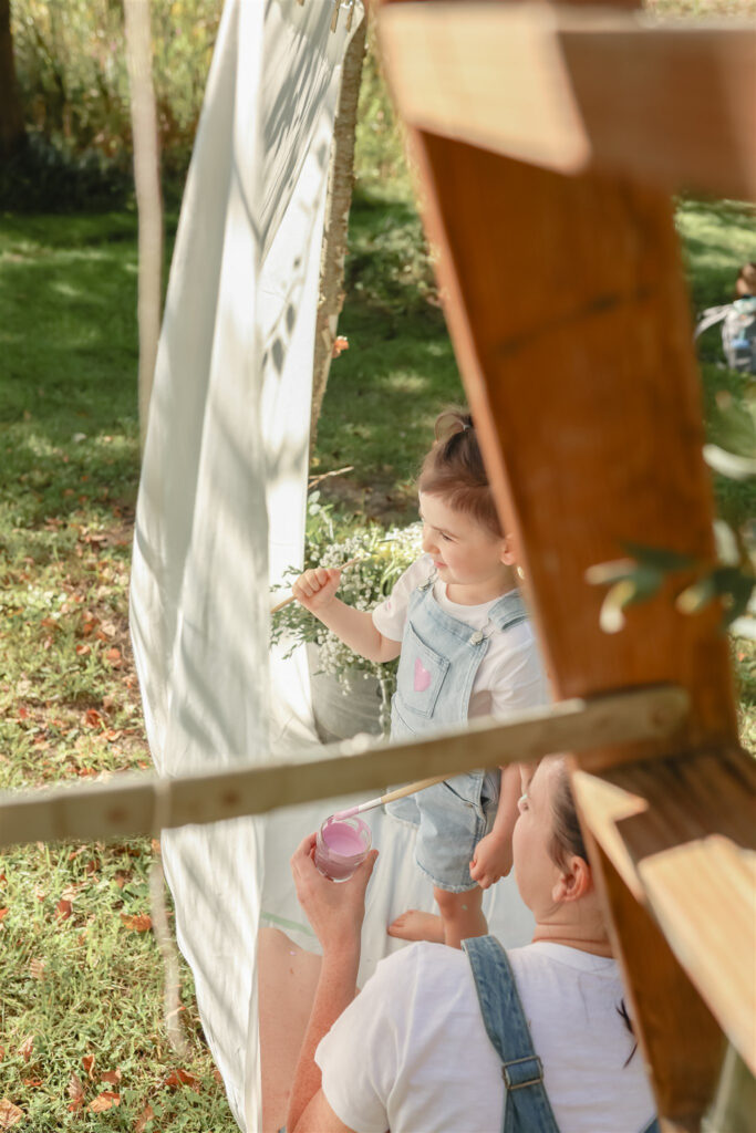 girl stands and paints on a sheet hanging between a tree and a wooden ladder during a mother daughter paint party