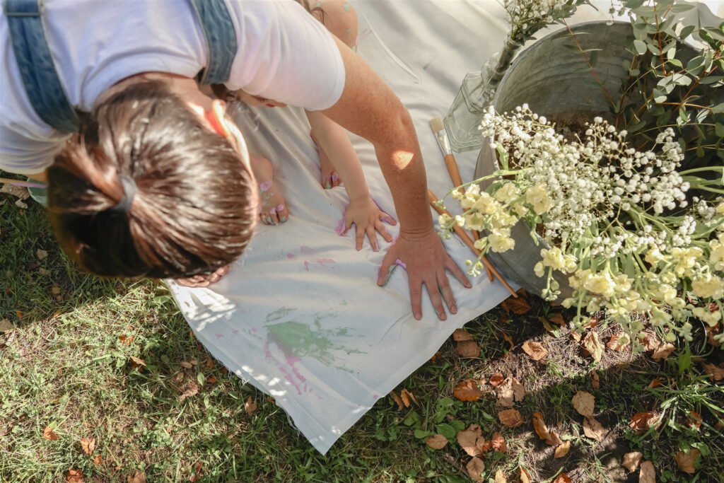 mother and daughter place painted hands down onto a sheet to make hand prints in this photo shot from above during a mother daughter paint party photoshoot