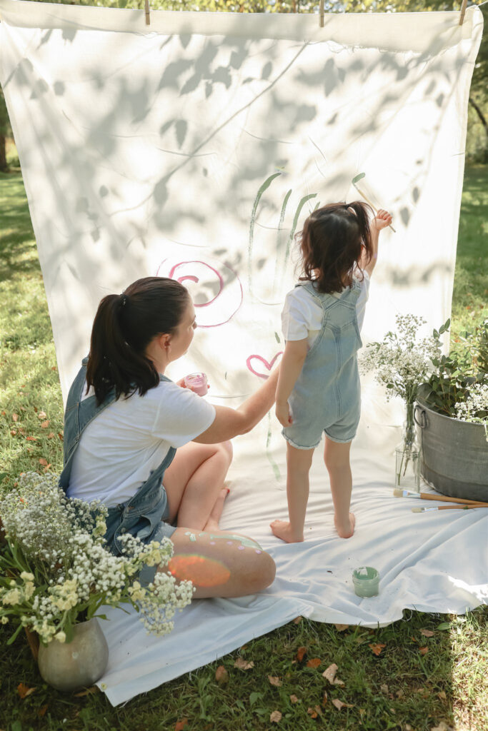 young girl and her mom paint on a sheet in a field surrounded by flowers in vases and buckets at a mother daughter paint party photoshoot