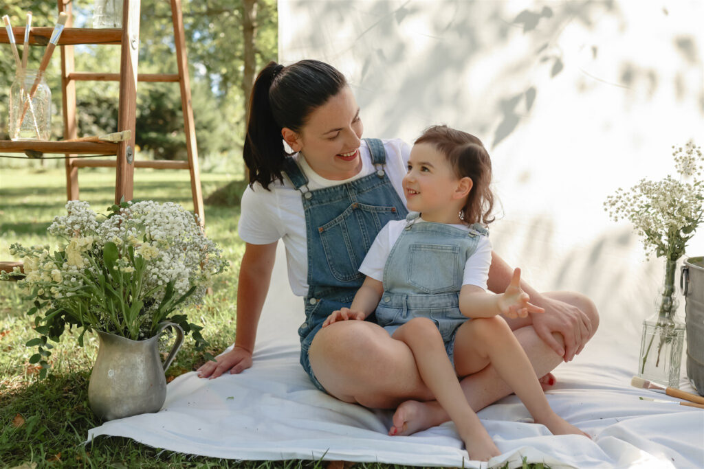 girl sits on mother's lap and they look at each other while surrounded by props for a mother daughter paint party photoshoot