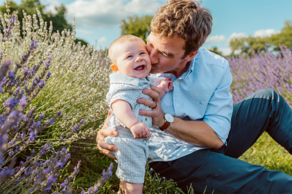father kisses baby son while sitting on the ground in between rows of purple and white lavender at lavender lane in milan michigan