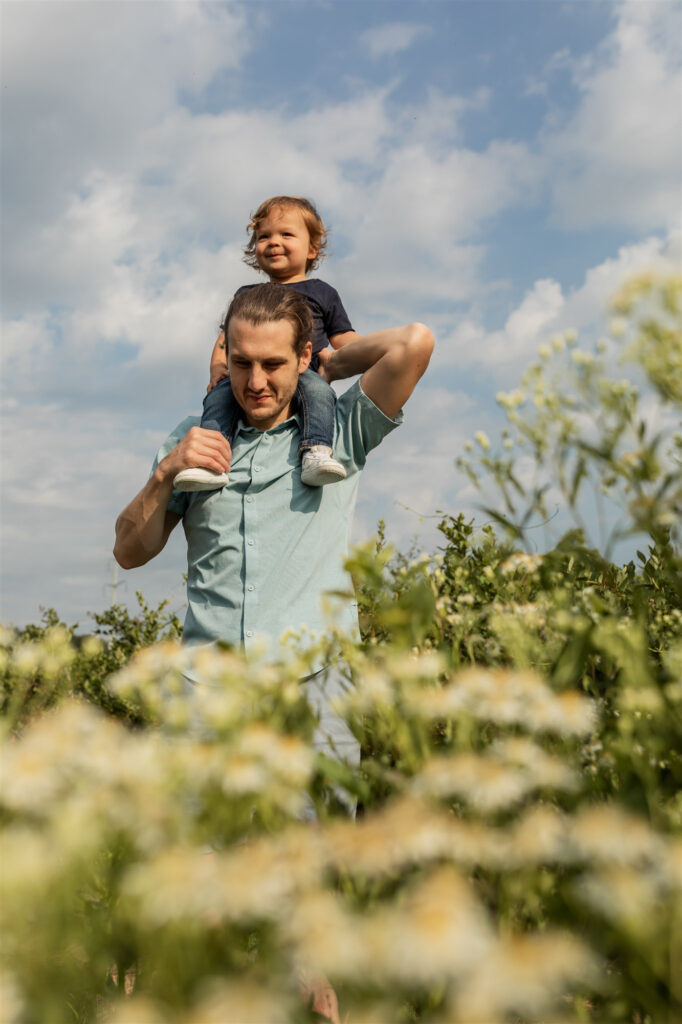 father gives shoulder ride for toddler son in photo shot from below with white flowers in the foreground
