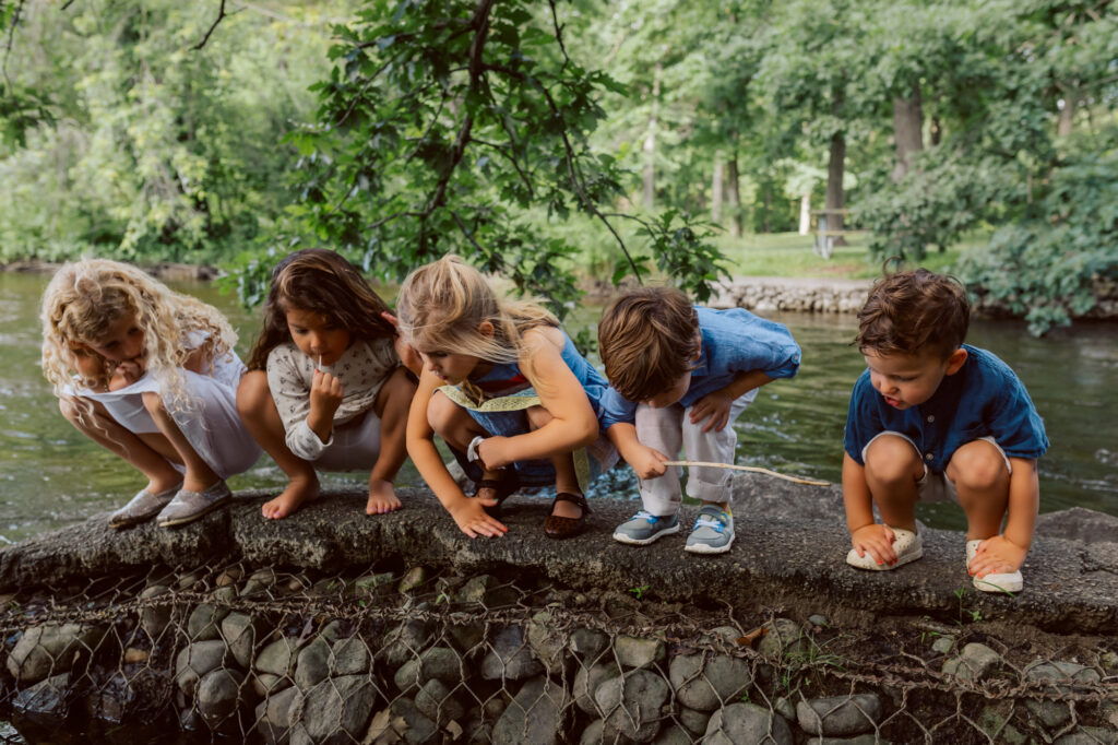 Group of children crouching along the riverbank, exploring nature together during an outdoor family photography session in Michigan
