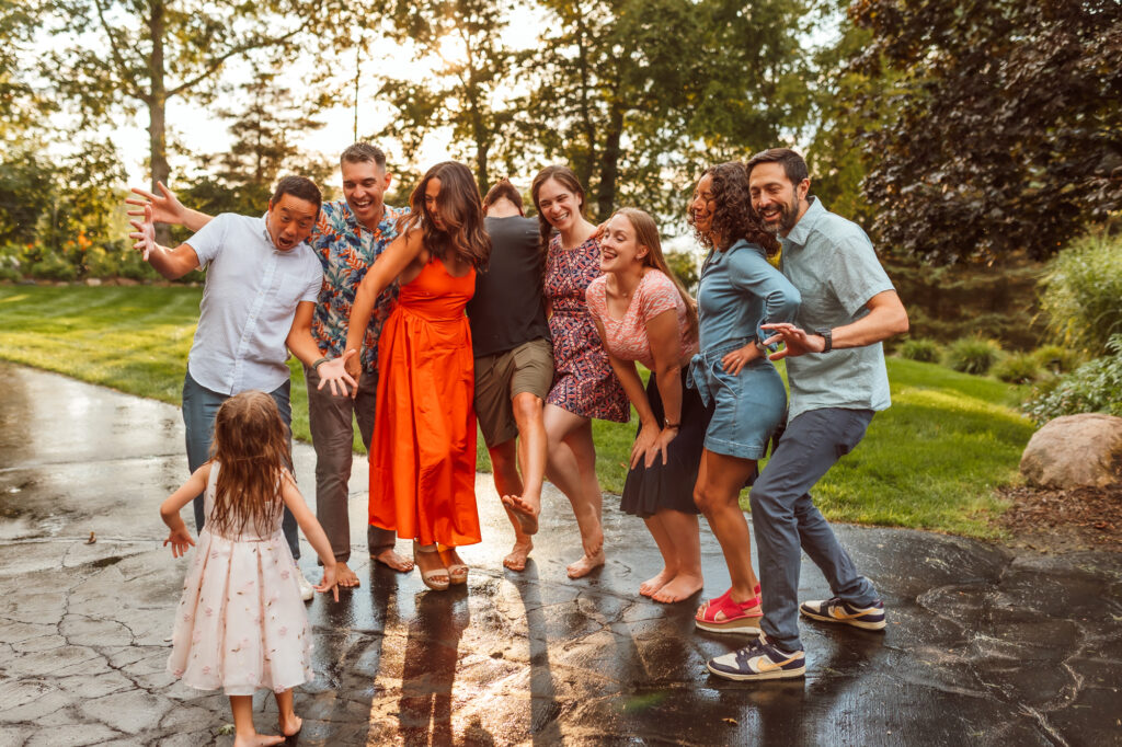 Extended family laughing and playing together outdoors after a summer rain, with a barefoot little girl in a white dress running toward the group, captured by Emmy & Ollie Photography, lifestyle family photographer