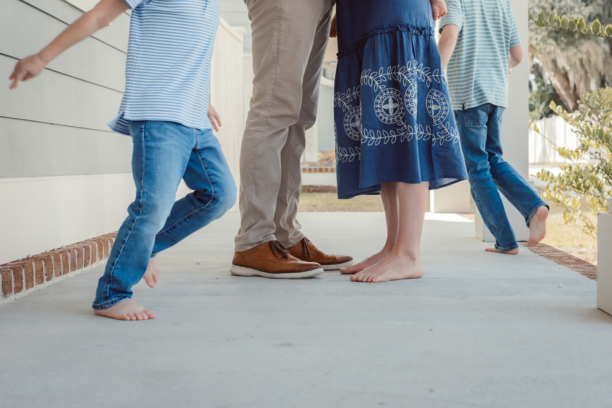 Playful family photography session by an Ann Arbor family photographer, capturing parents standing close together while their barefoot children run and play around them on the porch