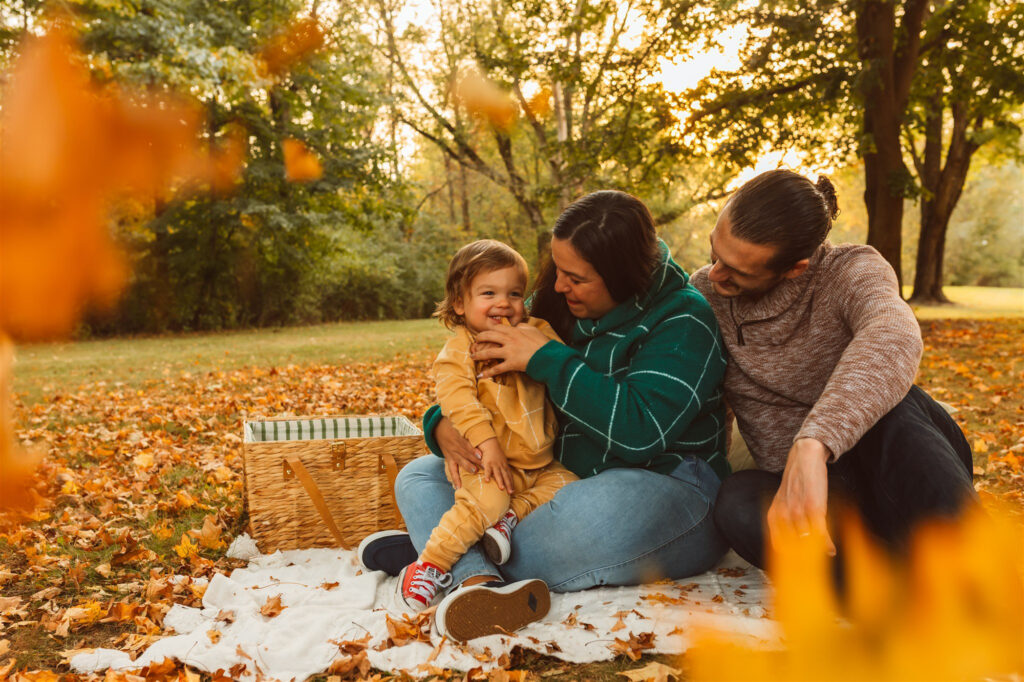 A family sits on a blanket surrounded by colorful autumn leaves at Wilderness Park in Saline, Michigan, smiling and cuddling during a fall family photo session by Emmy & Ollie Photography