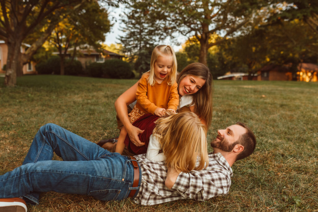 A playful family of four laughs together on the grass during a fall family photo session at Virginia Park in Ann Arbor, Michigan, captured by Emmy & Ollie Photography with warm golden-hour light and candid smiles