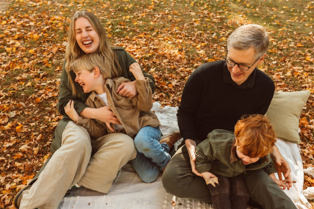 A joyful family of four laughs and plays together on a blanket covered in fall leaves during a cozy autumn photo session at Wilderness Park in Saline, Michigan, captured by Emmy & Ollie Photography