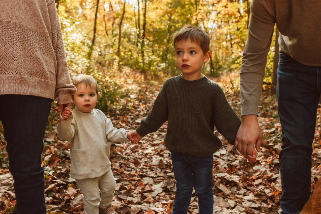 Two young children hold hands with their parents while walking along a wooded trail covered in fall leaves at Wilderness Park in Saline, Michigan, during a warm and natural family photo session by Emmy & Ollie Photography