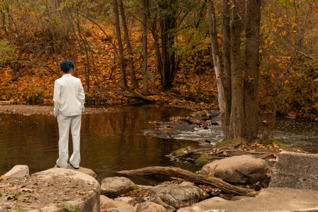 A person in a white suit stands on rocks beside the river at Parker Mill County Park in Ann Arbor, Michigan, surrounded by golden fall leaves and trees during an autumn photo session captured by Emmy & Ollie Photography