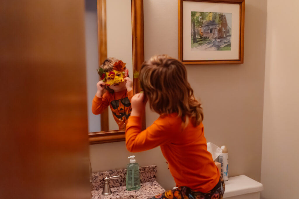 A young boy looks at himself in the bathroom mirror while wearing a handmade fall mask during a cozy family craft night, captured as part of an in-home documentary style family photo session in Saline, Michigan