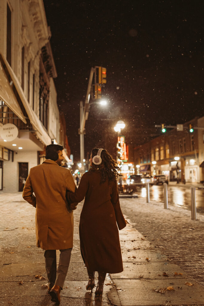 A couple walks arm in arm down a quiet street during a gentle snowfall in downtown Ann Arbor, Michigan, bundled in winter coats and sharing an intimate moment during a documentary style couples photo session.