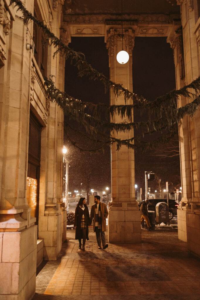 A couple strolls together beneath the historic arches of Nickels Arcade in Ann Arbor, Michigan, as light snow falls during a cozy winter couples photo session captured in a natural, documentary style.