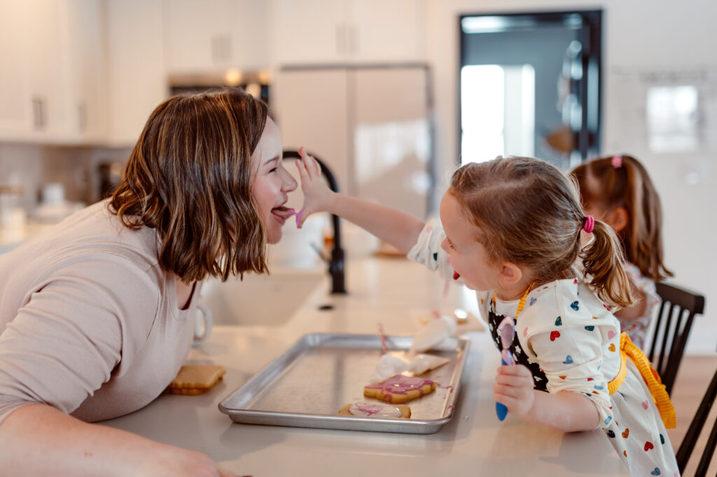 A young child playfully feeds frosting to her mom while decorating cookies during a silly in-home documentary style baking session, capturing real laughter and connection in an Ann Arbor, Michigan family photo shoot by a family photographer.