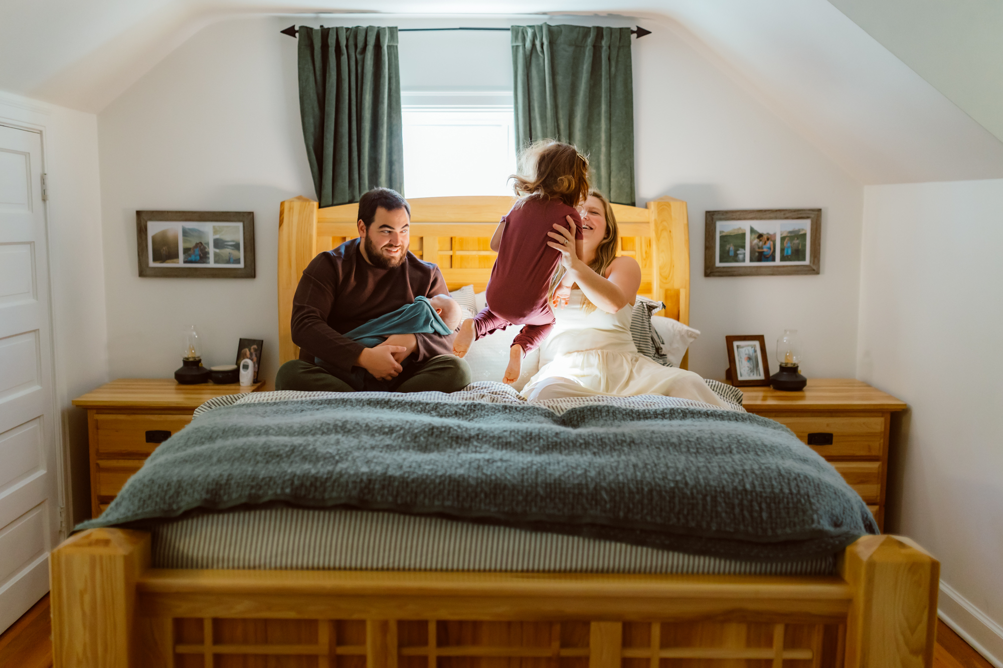 A family sits together on the bed during an in-home newborn photo session in Ann Arbor, Michigan, with parents cuddling their newborn while a toddler jumps and plays nearby, captured by a Michigan lifestyle newborn photographer.