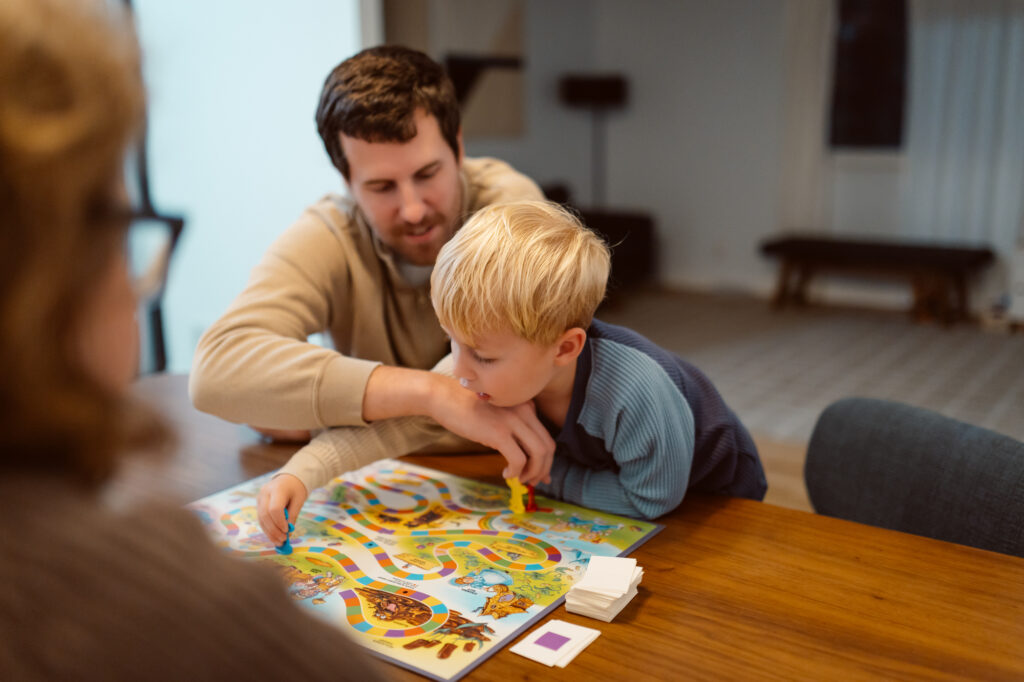 A young boy plays Candy Land with his parents at the dining table during a cozy in-home documentary style family photo session in Saline, Michigan, captured by a Michigan lifestyle family photographer.