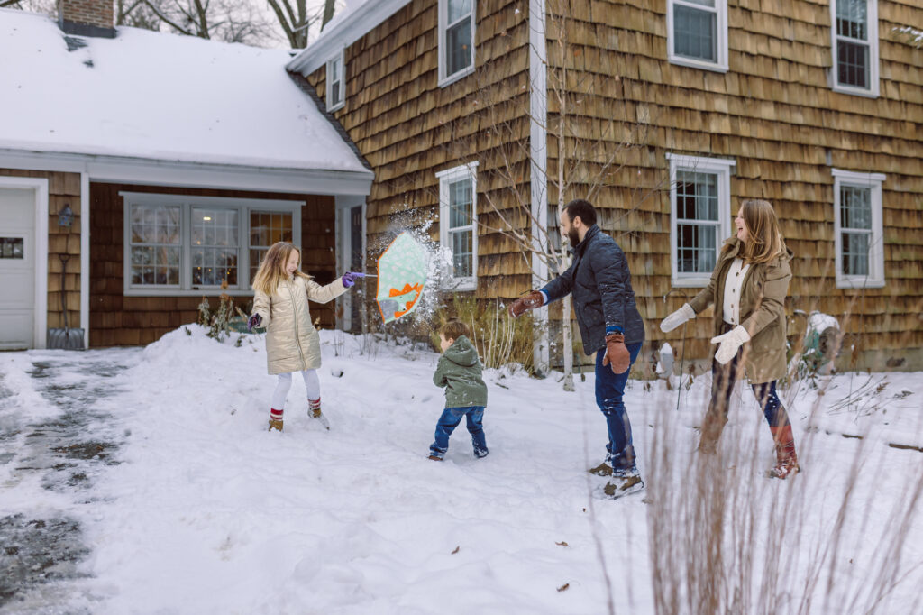A family laughs and plays together during a playful snowball fight in their front yard during an at-home documentary style family photo session in Michigan, capturing candid winter joy and real connection