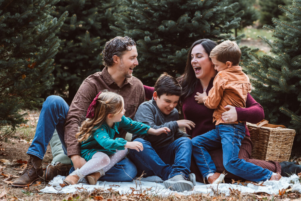 A playful family of five laughs together while sitting on a blanket among evergreen trees during an outdoor family photo session in Dexter, Michigan at Westman's Tree Farm