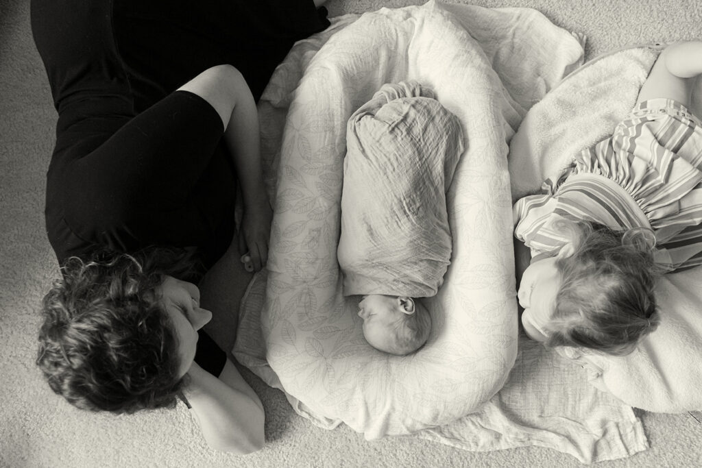 A mother and her older child laugh and cuddle on the floor beside a swaddled newborn during a lifestyle newborn photo session in Ann Arbor, Michigan, photographed by a Michigan family and newborn photographer.