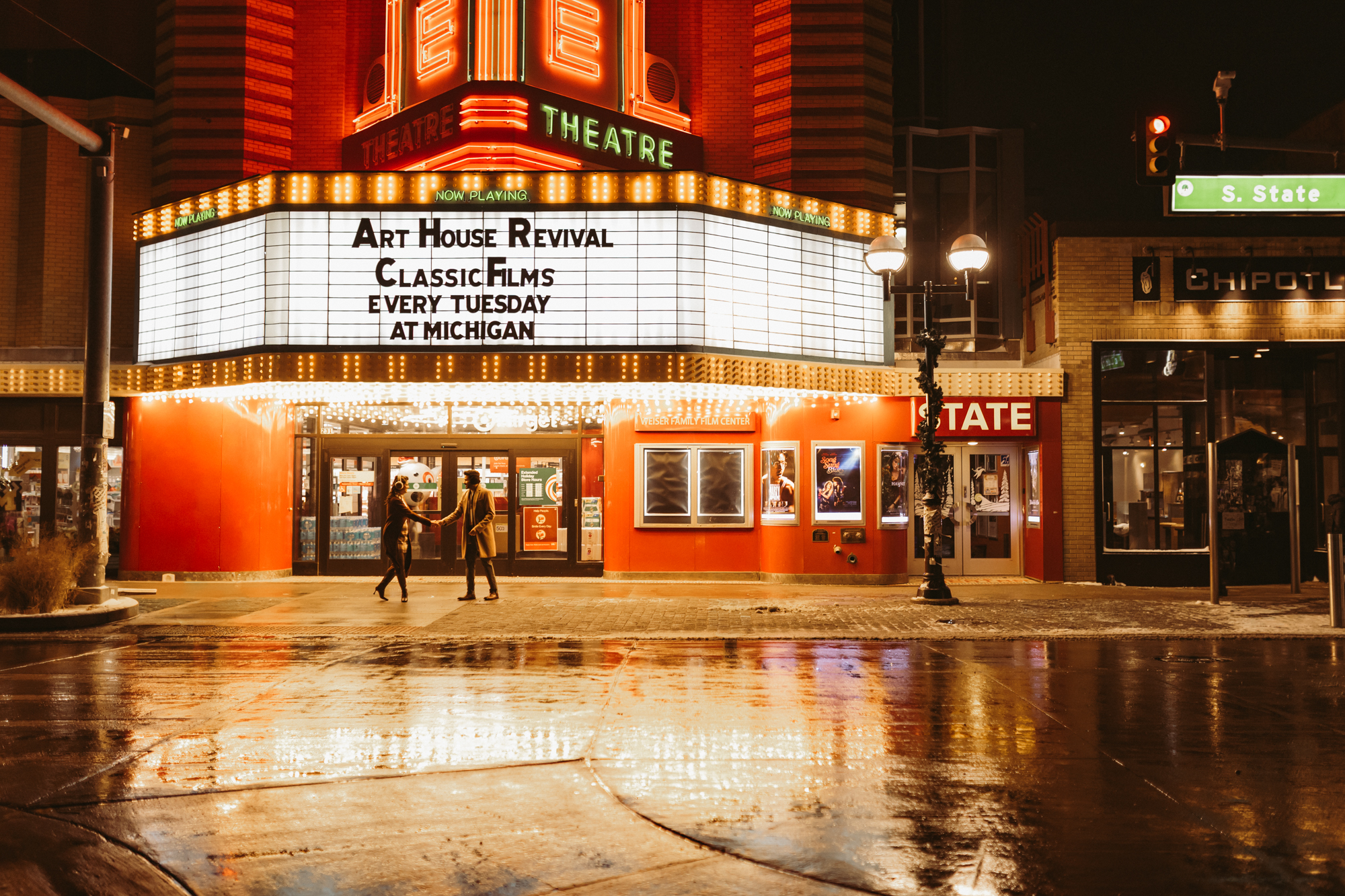 A couple dances together outside the State Theatre in downtown Ann Arbor, Michigan, surrounded by glowing marquee lights and fresh snow during a romantic winter couples photo session with a lifestyle photographer.