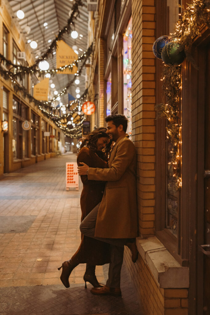 man and woman cuddle while leaning against wall in nickels arcade