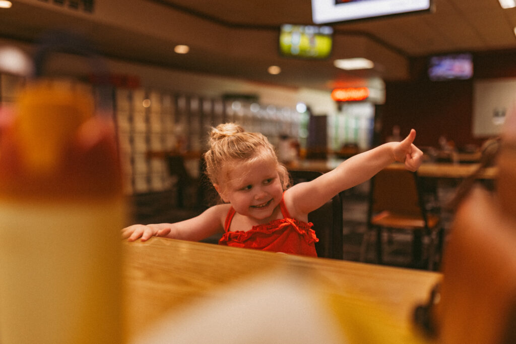 girl sits at a wooden table in a bowling alley and gives thumbs up to her brother