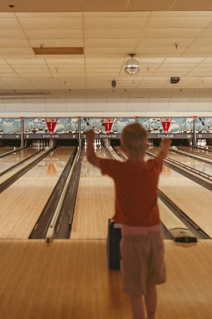 boy stands and watches bowling ball roll down lane while throwing up arms in cheer